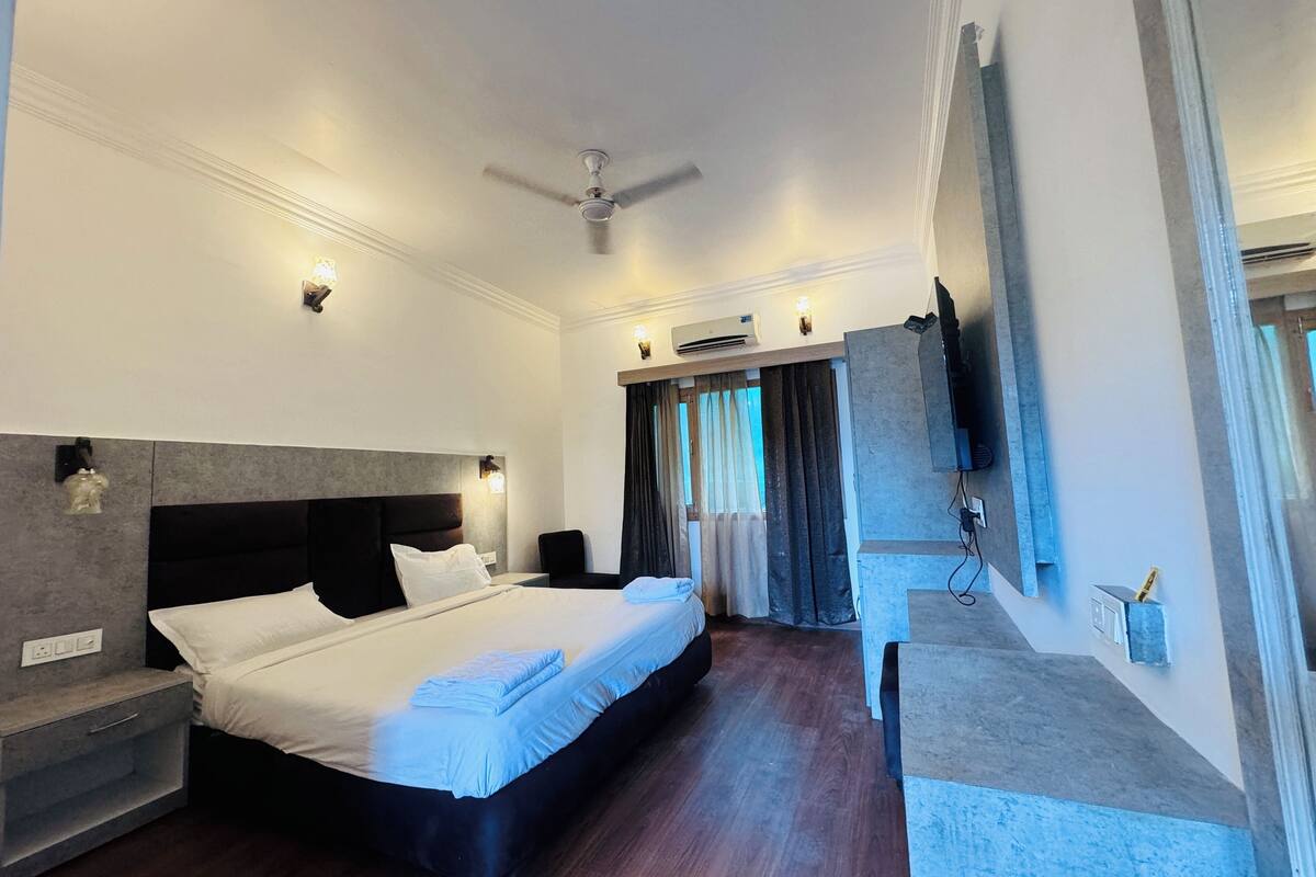 Quarto de hotel moderno com uma cama grande, um ventilador de teto, televisão de tela plana na parede e um pequeno espaço de estar com uma cadeira e uma mesa.