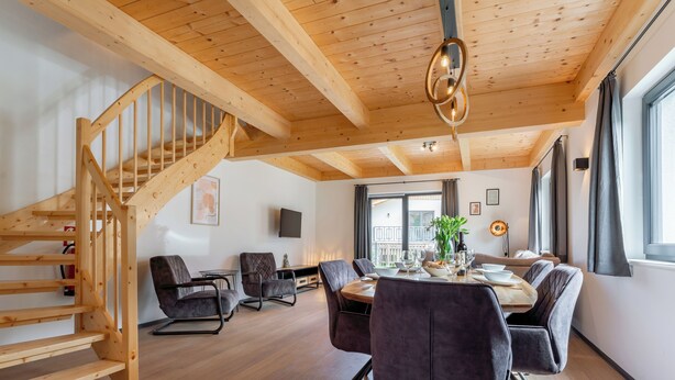 Un interior moderno con una escalera de madera, un comedor con una mesa redonda y sillas, y un gran ventanal que proporciona luz natural.