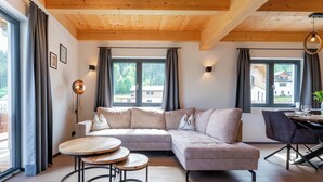 Una sala de estar moderna con un sofá seccional, mesas redondas de madera y ventanas amplias que ofrecen vistas al exterior.