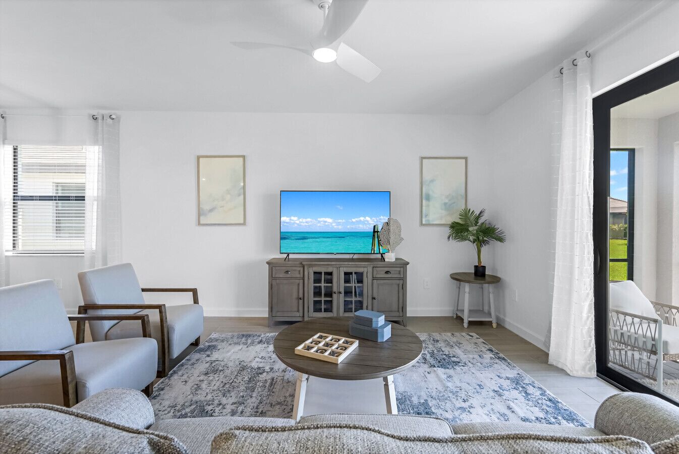 A living room with a flat-screen TV, a ceiling fan, a coffee table, and a view of the ocean through a large window.