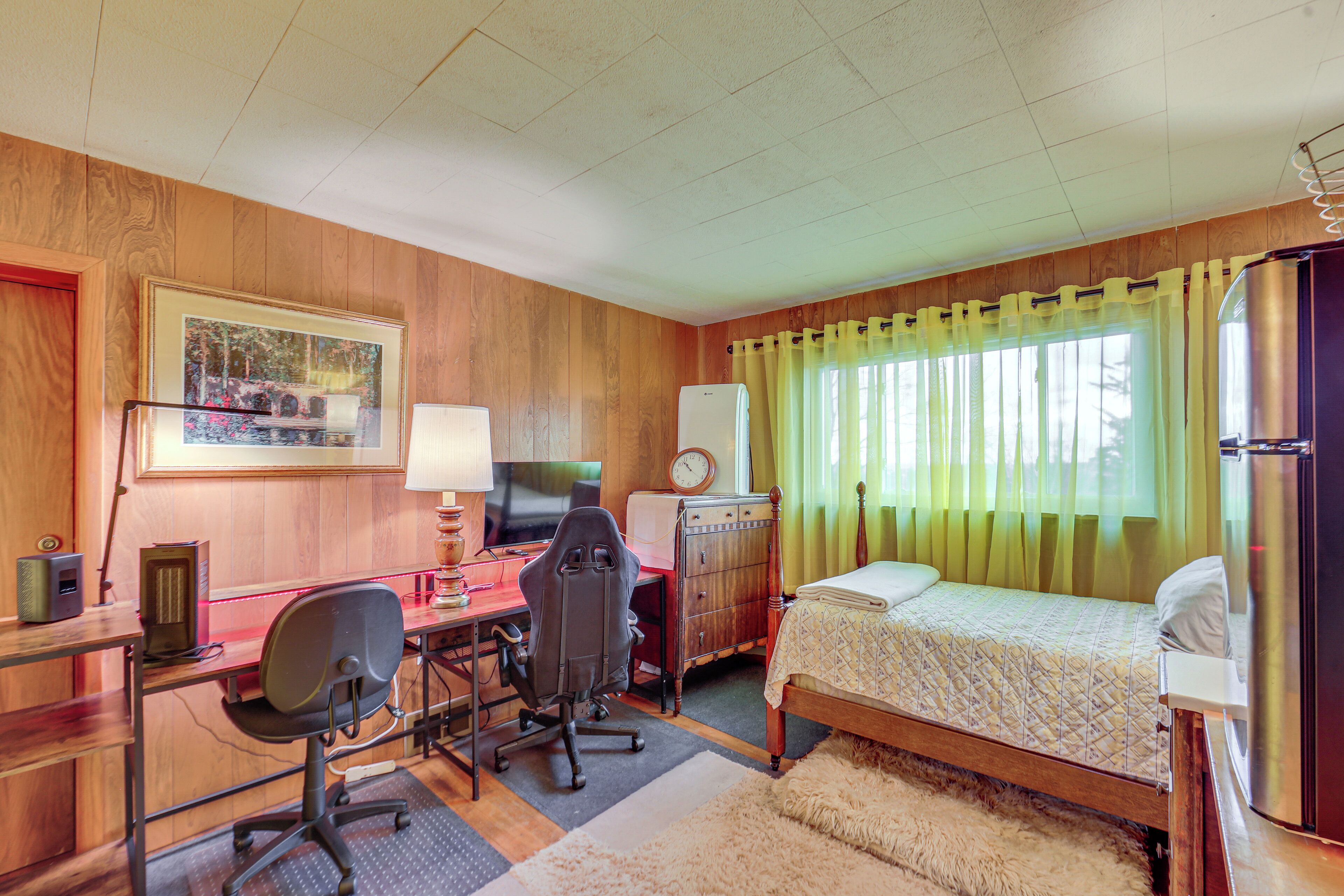 A room with a wooden paneled wall, a bed with a quilted bedspread, a desk with a computer and chair, a radiator, and a window with yellow curtains.