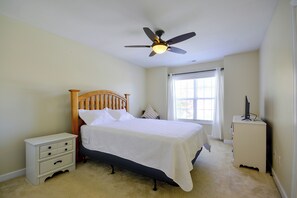 Quarto com cama, mesa de cabeceira, cômoda, ventilador de teto e janela com cortinas.