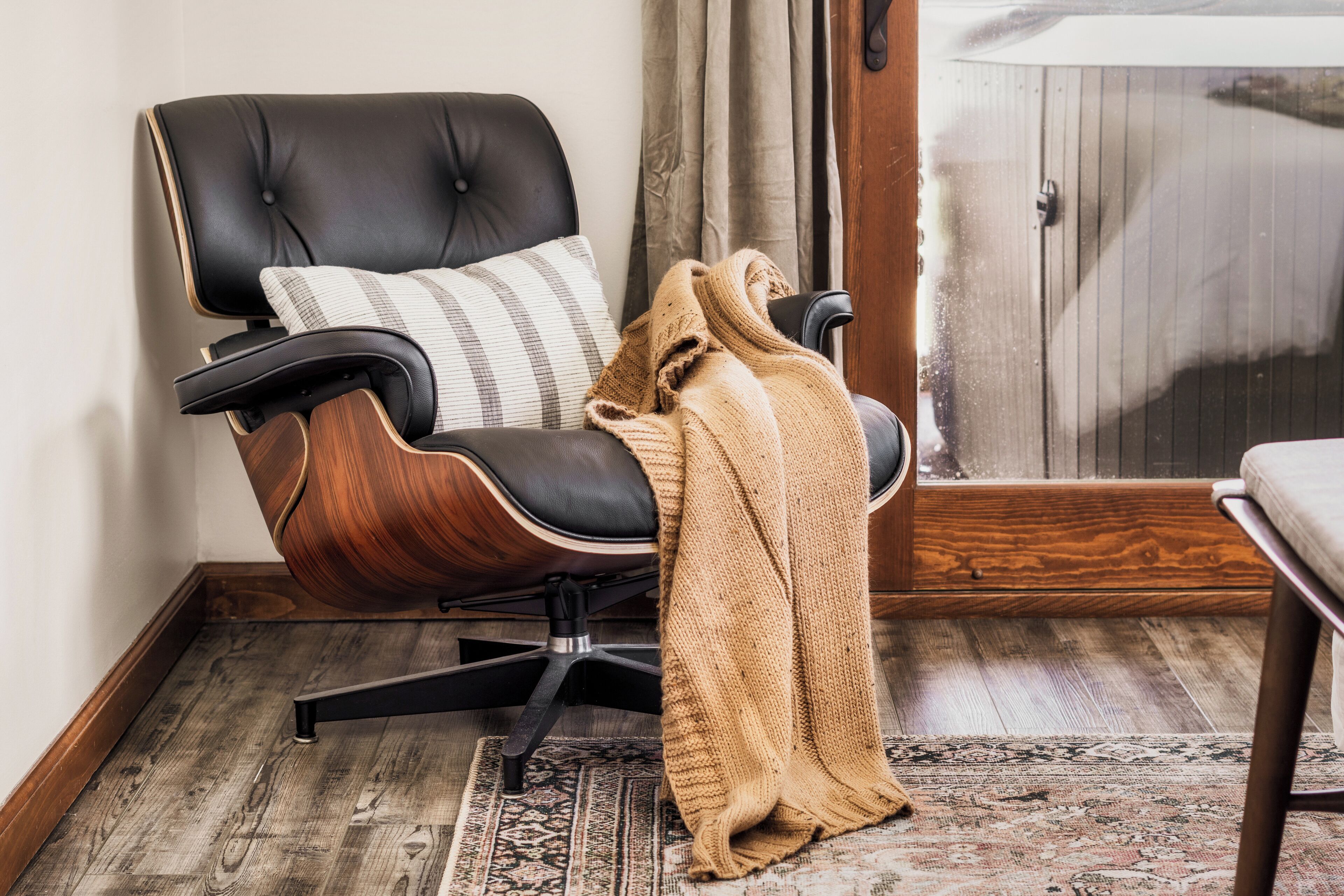 A modern chair with a wooden frame and black upholstery, a patterned cushion, and a knitted throw on the floor.