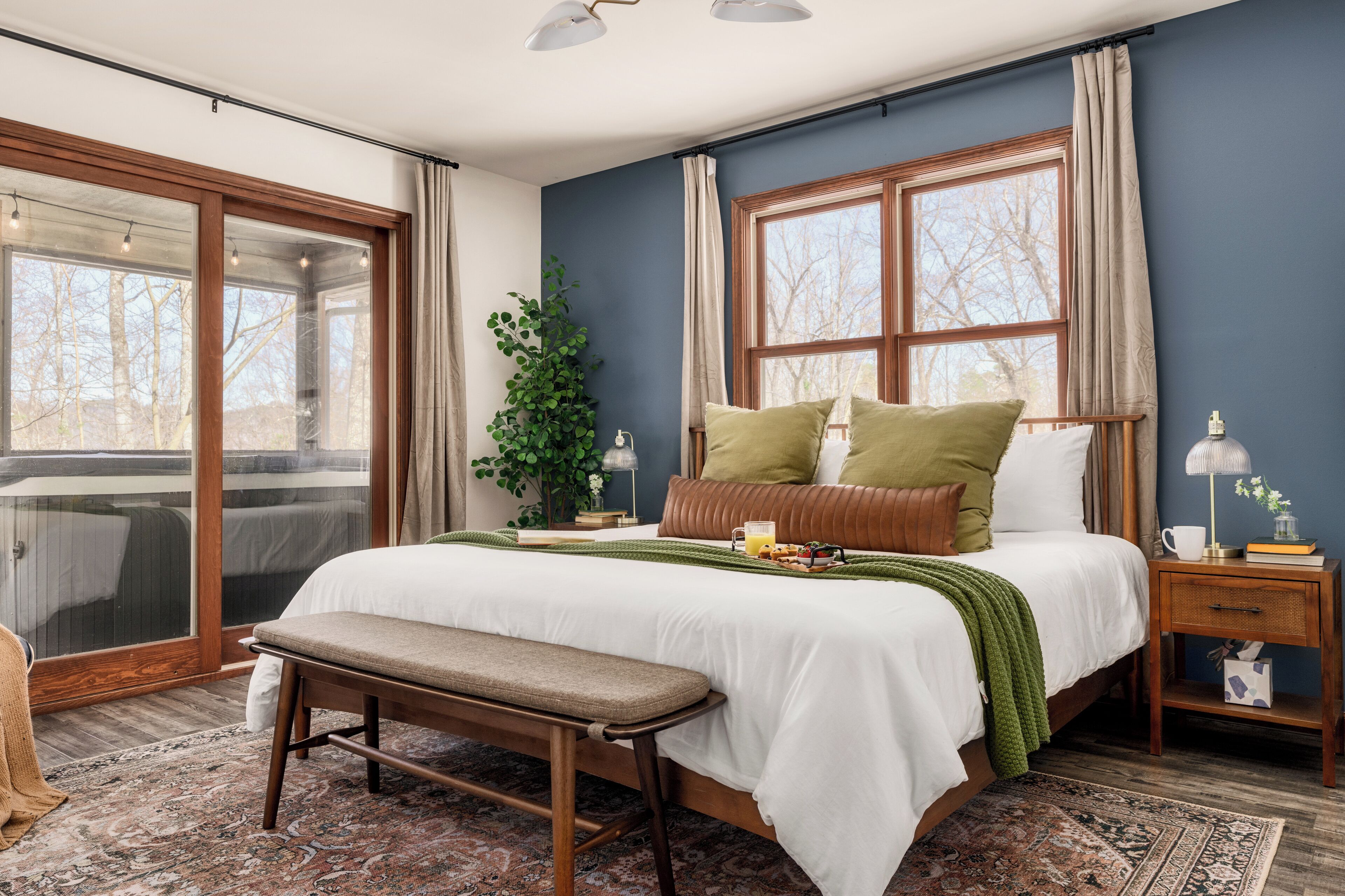 A bedroom with a large bed, a bench, a nightstand, a lamp, and a window with curtains.