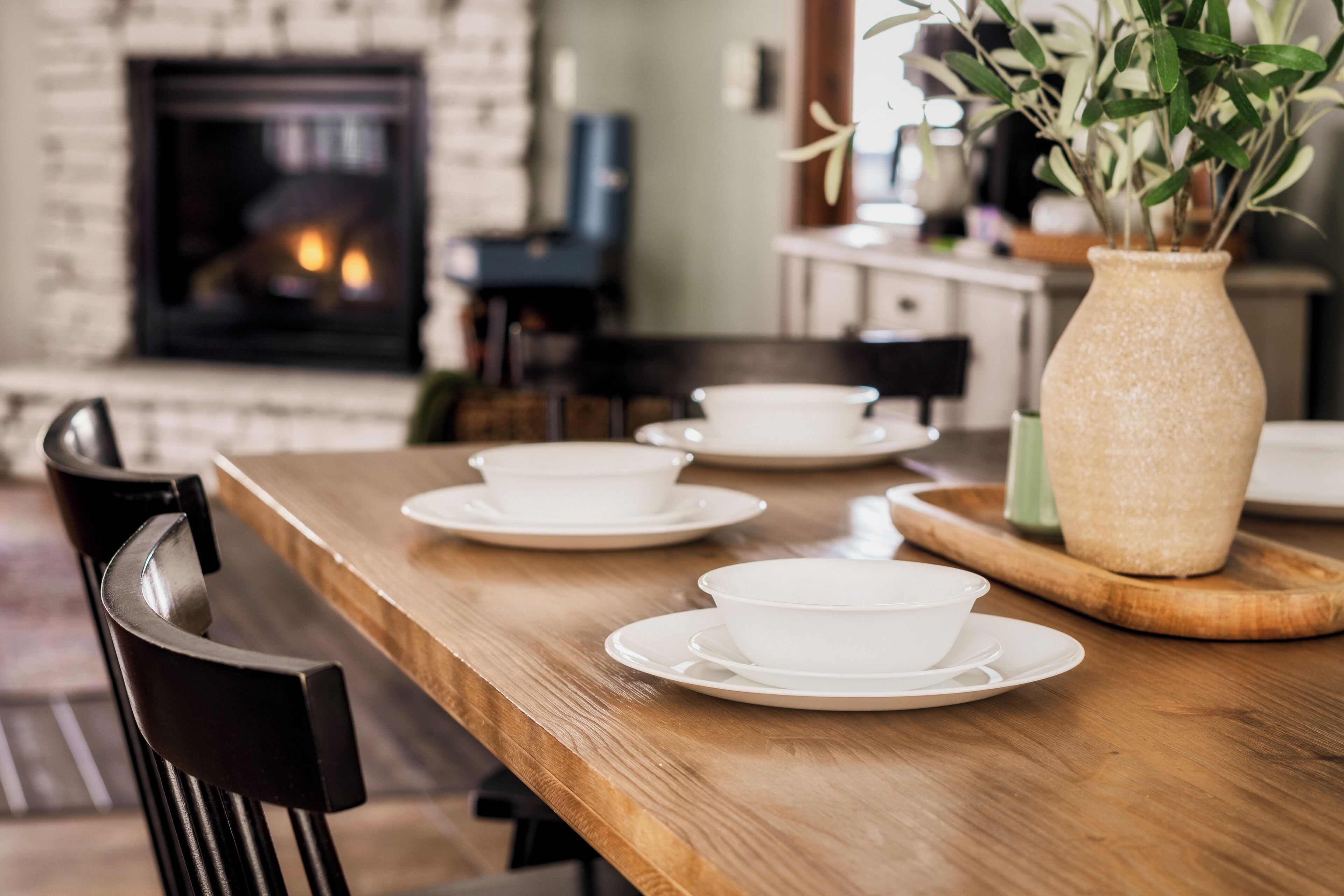 A dining area with a wooden table set with white bowls and saucers, a vase with greenery, and a brick fireplace in the background.