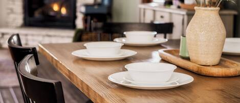A dining area with a wooden table set with white bowls and saucers, a vase with greenery, and a brick fireplace in the background.