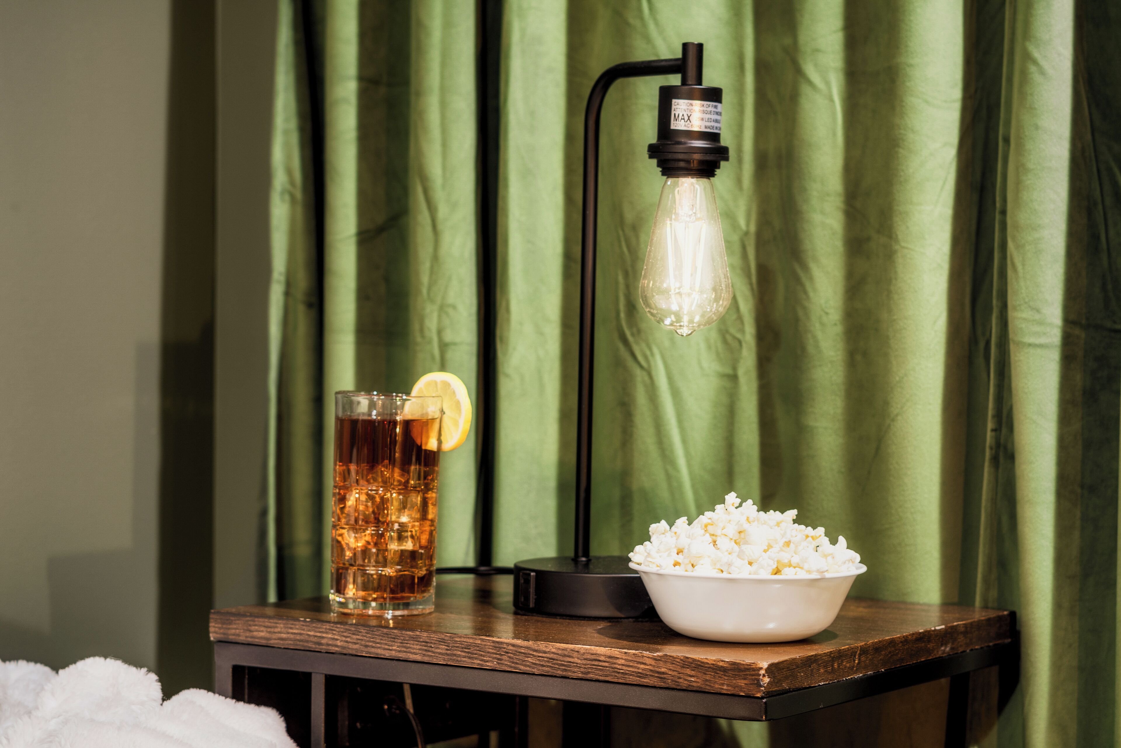 A table with a glass of iced tea, a bowl of popcorn, and a table lamp against a green curtain backdrop.