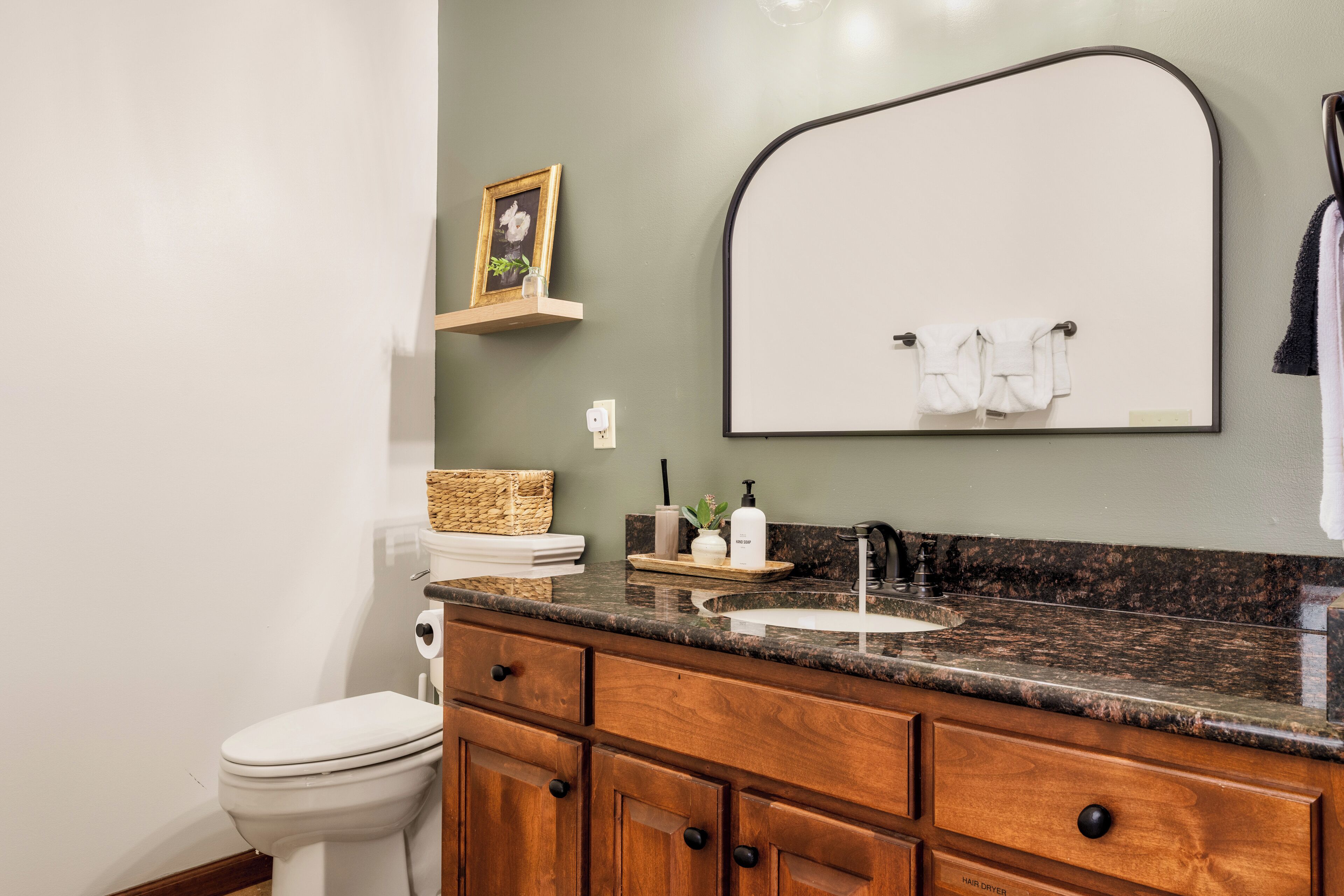 A bathroom with a toilet, wooden vanity, granite countertop, sink, and a mirror with a towel rack.