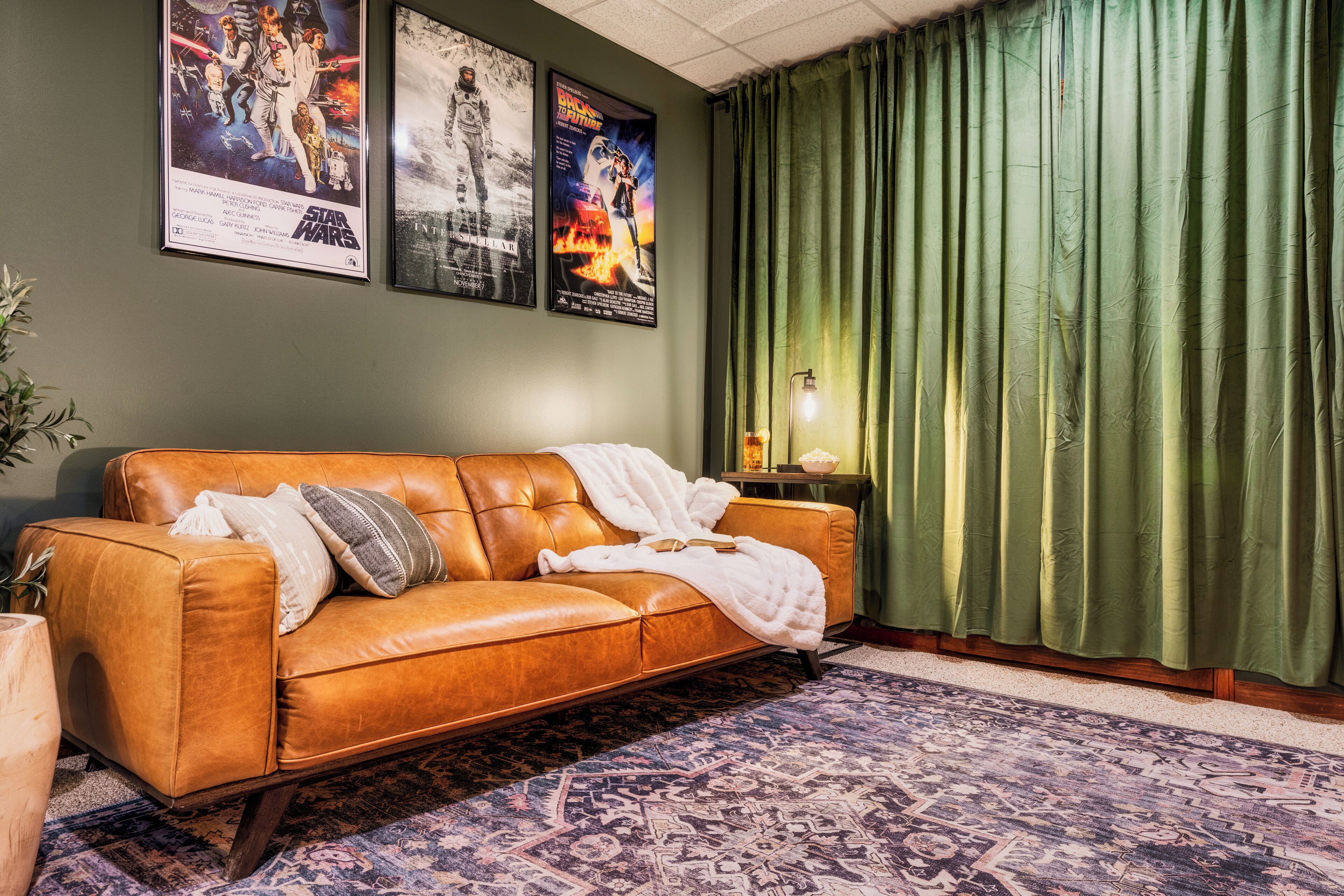 A room with a brown leather sofa, a patterned rug, green curtains, and two movie posters on the wall.