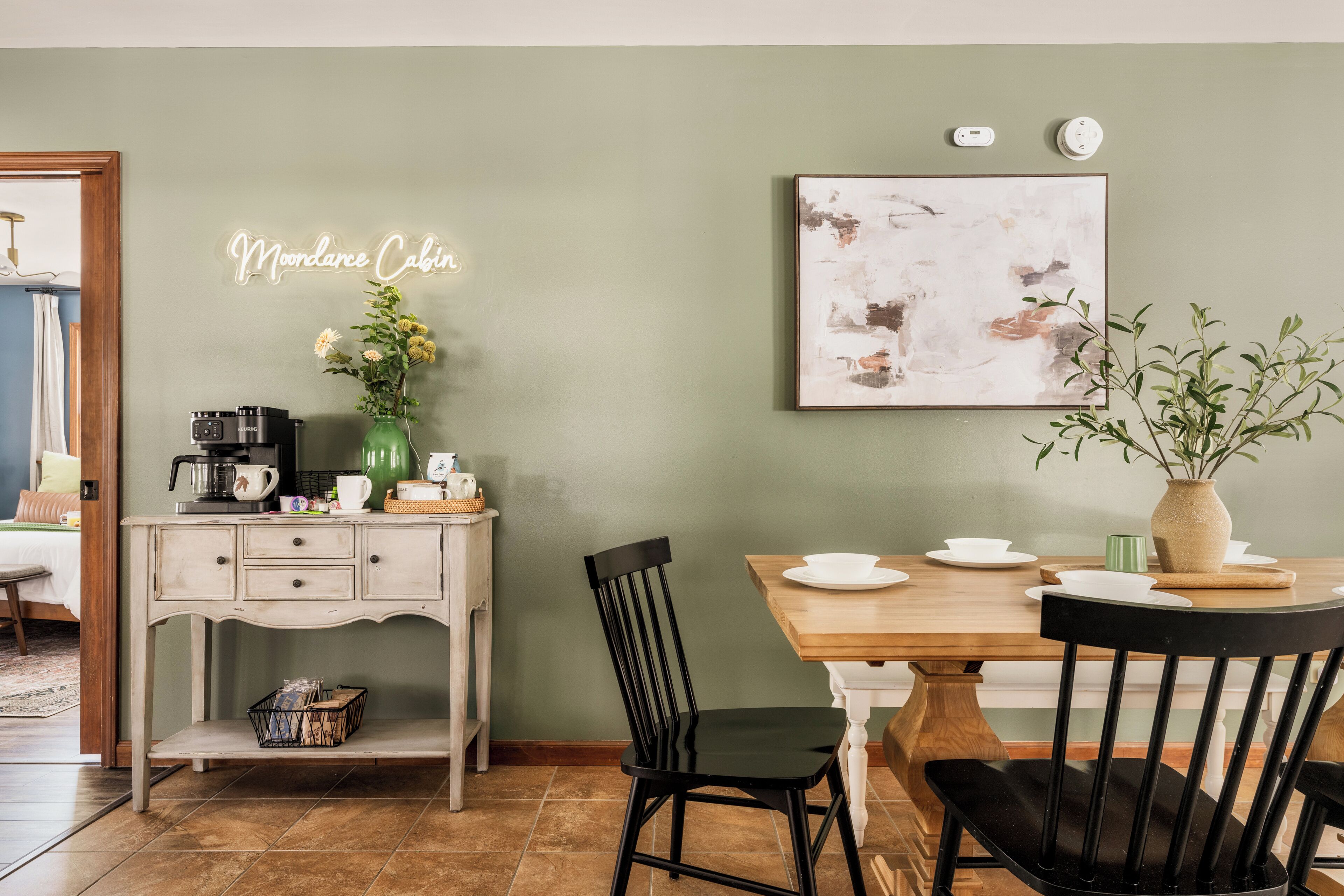 Un comedor con mesa de madera, sillas negras, una cafetera y un florero con flores.