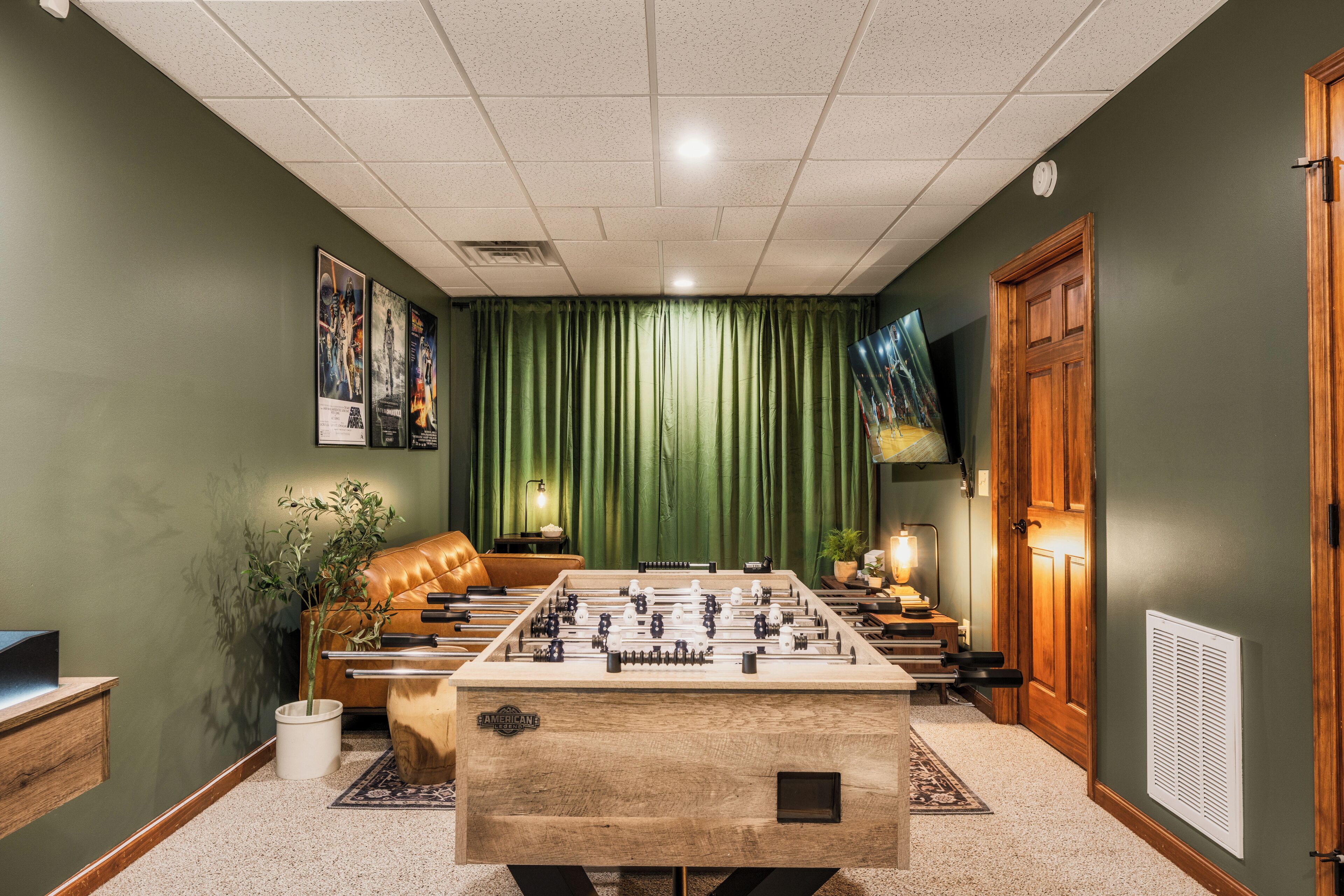 A room with a foosball table, a leather couch, a television, and a green curtain.