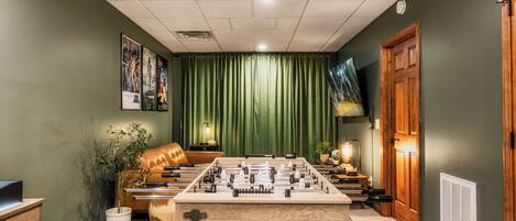 A room with a foosball table, a leather couch, a television, and a green curtain.