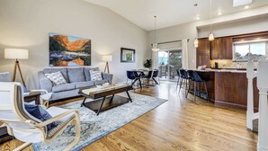A spacious living room with a sofa, coffee table, and chairs, a dining area with a table and chairs, a kitchen with wooden cabinets and a bar, and a large window with a view.