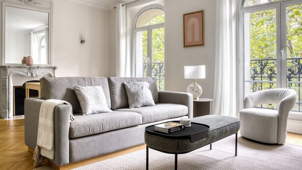 A living room with a grey sofa, a round coffee table, and a grey armchair. There is a fireplace, a lamp, and a large window with a view of trees. - Blueground | Porte Saint Cloud, concierge (Paris)