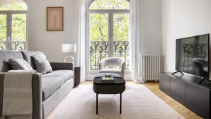A modern living room with a grey sofa, a small black coffee table, a white armchair, and a TV on a black cabinet. - Blueground | Porte Saint Cloud, concierge (Paris)