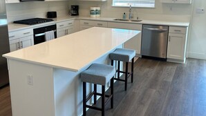 A modern kitchen with white cabinetry, a large island with bar stools, a microwave, a coffee maker, and a dishwasher. - NEW Smart Home w/ Massage Chair & King Bed (Lathrop)