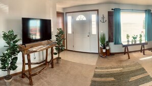 A living room with a flat-screen TV on a wooden stand, a large window with curtains, and several potted plants. - Homestead Stays, Heart of the area's top attractions (Oak Harbor)