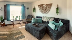 A living room with a sectional sofa, a coffee table, and a rug. There is a window with curtains, a map on the wall, and a plant on the windowsill. - Homestead Stays, Heart of the area's top attractions (Oak Harbor)