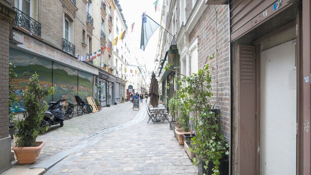 Une ruelle étroite pavée de pierres, bordée de bâtiments des deux côtés, dont certains ont les volets fermés, et quelques plantes en pot le long du chemin.