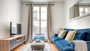 Un salon moderne avec un canapé bleu, un téléviseur à écran plat, une console en bois et une fenêtre avec des rideaux.