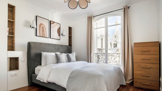 Ein Schlafzimmer mit Bett, einem Holzkleiderschrank, einem Balkon mit Blick auf Gebäude und einer Wand mit gerahmten Kunstwerken.