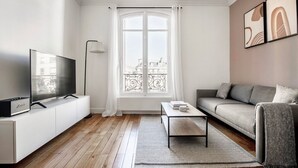 Ein modernes Wohnzimmer mit einem Flachbildfernseher, einem grauen Sofa, einem Couchtisch und einem großen Fenster mit Blick auf Gebäude.