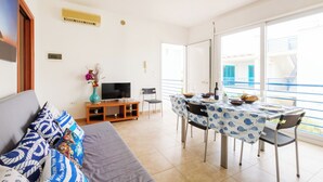 Ein Wohnzimmer mit Sofa, Fernseher und Esstisch mit Stühlen. Ein Balkon mit Geländer und Blick auf Gebäude ist vorhanden.