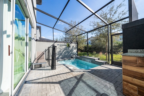 A modern outdoor swimming pool area with a glass enclosure, a brick patio, and a wooden deck.