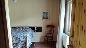 Une chambre à coucher avec un lit, une chaise, un ventilateur de plafond, une fenêtre avec des rideaux et un plancher en bois.