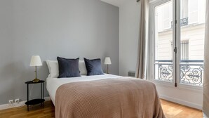 A bedroom with a bed, two bedside lamps, a window with a view of buildings, and a wooden floor. - Blueground | Champs Elysees, nr Arc de Triomphe (Paris)