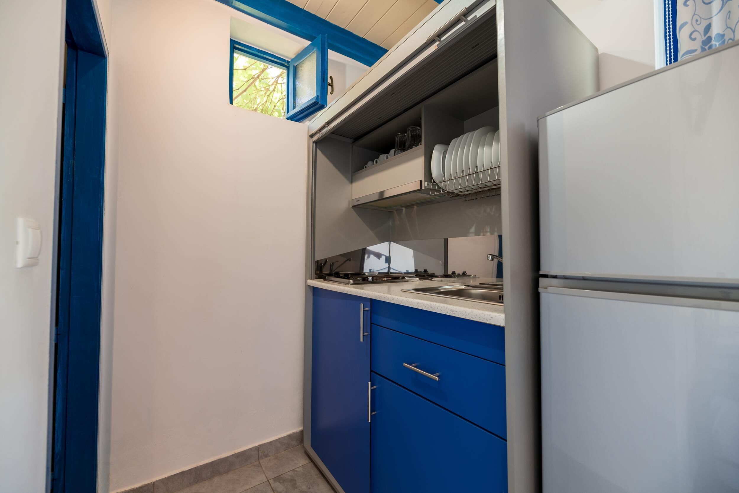 A compact kitchen with blue cabinets, a stainless steel refrigerator, and a window above the sink.
