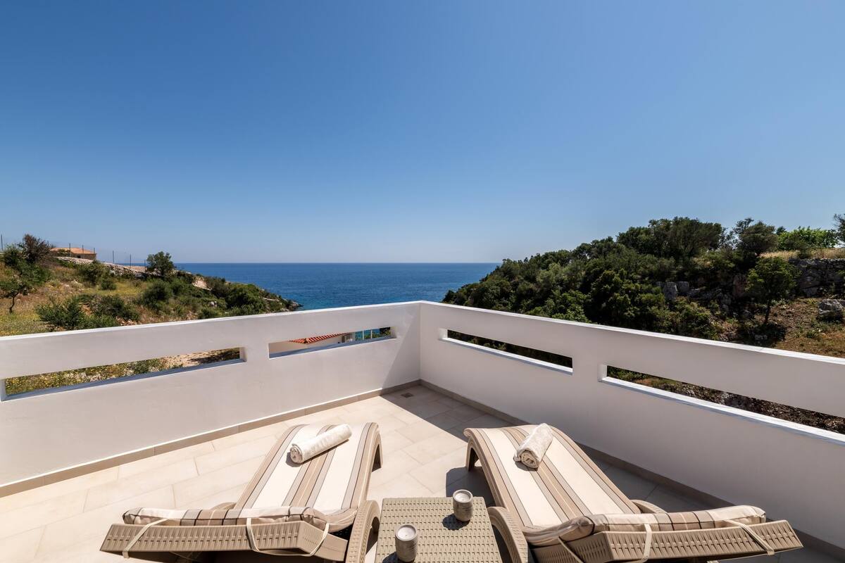 A balcony with two sun loungers and a table, overlooking a coastal view.