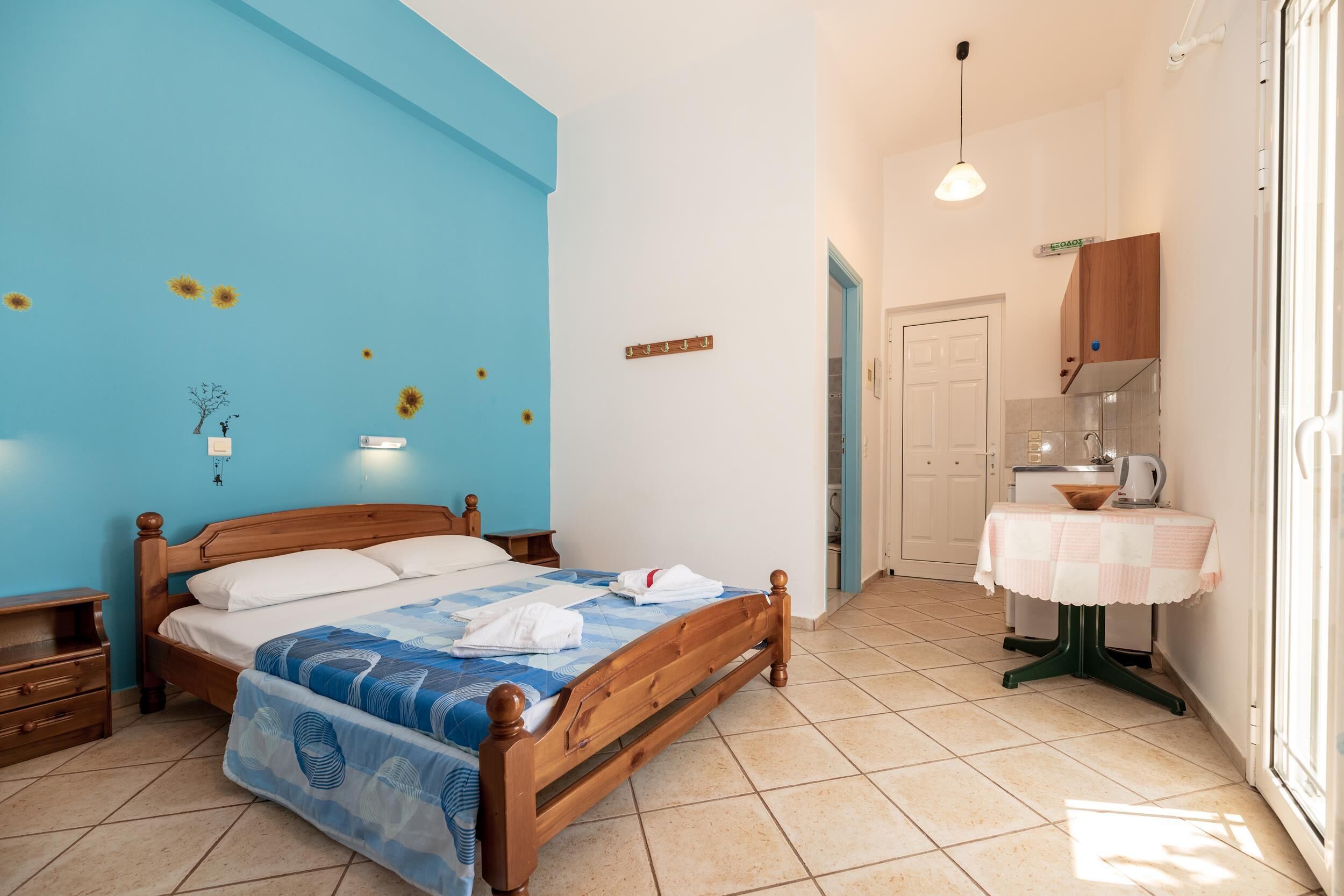 A bedroom with a wooden bed, a small bedside table, a nightstand, a wall-mounted light, a bathroom with a sink and mirror, and a window with a view.