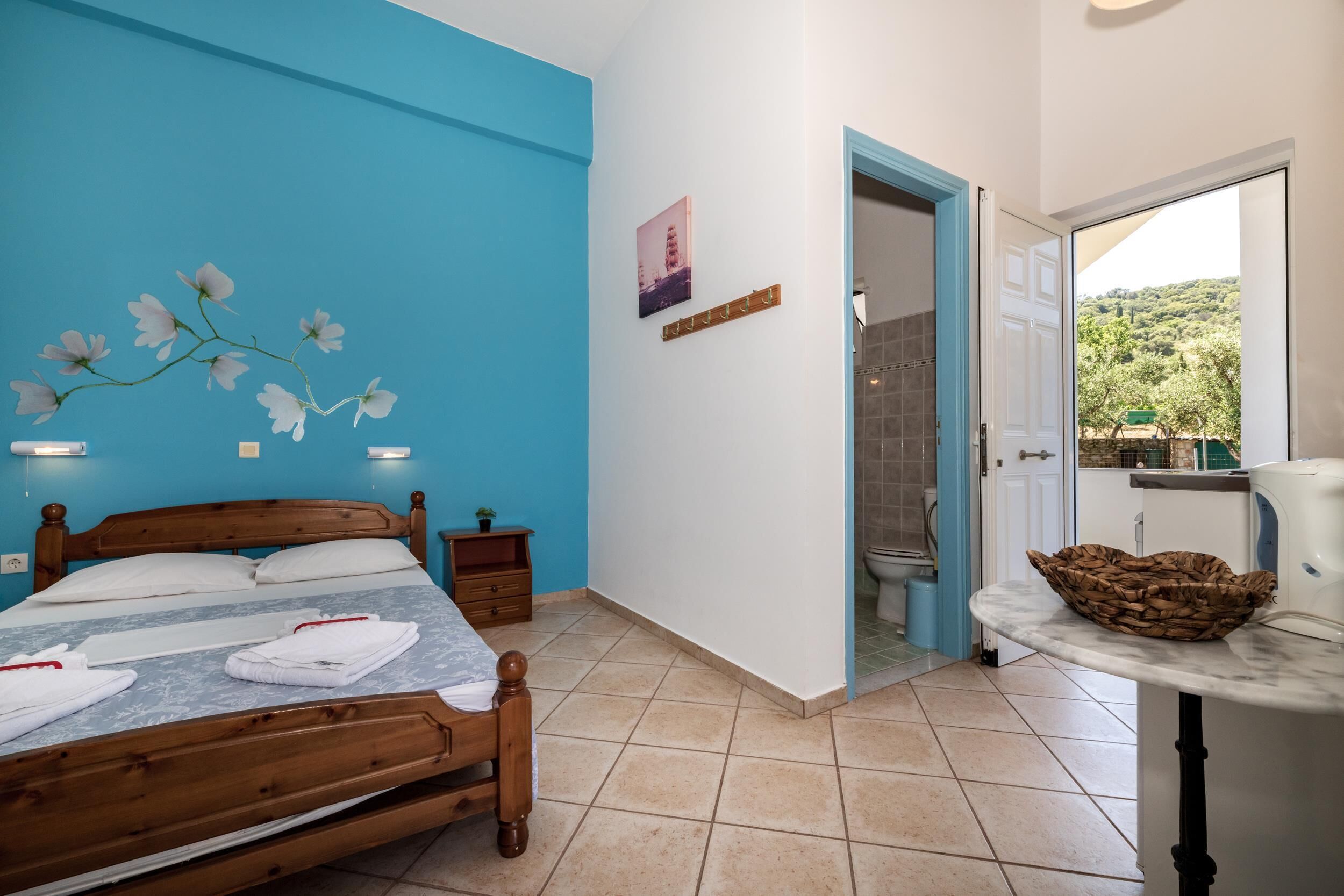 A bedroom with a wooden bed, a blue accent wall, a small bedside table, a nightstand, a bathroom visible through an open door, and a view of greenery outside.