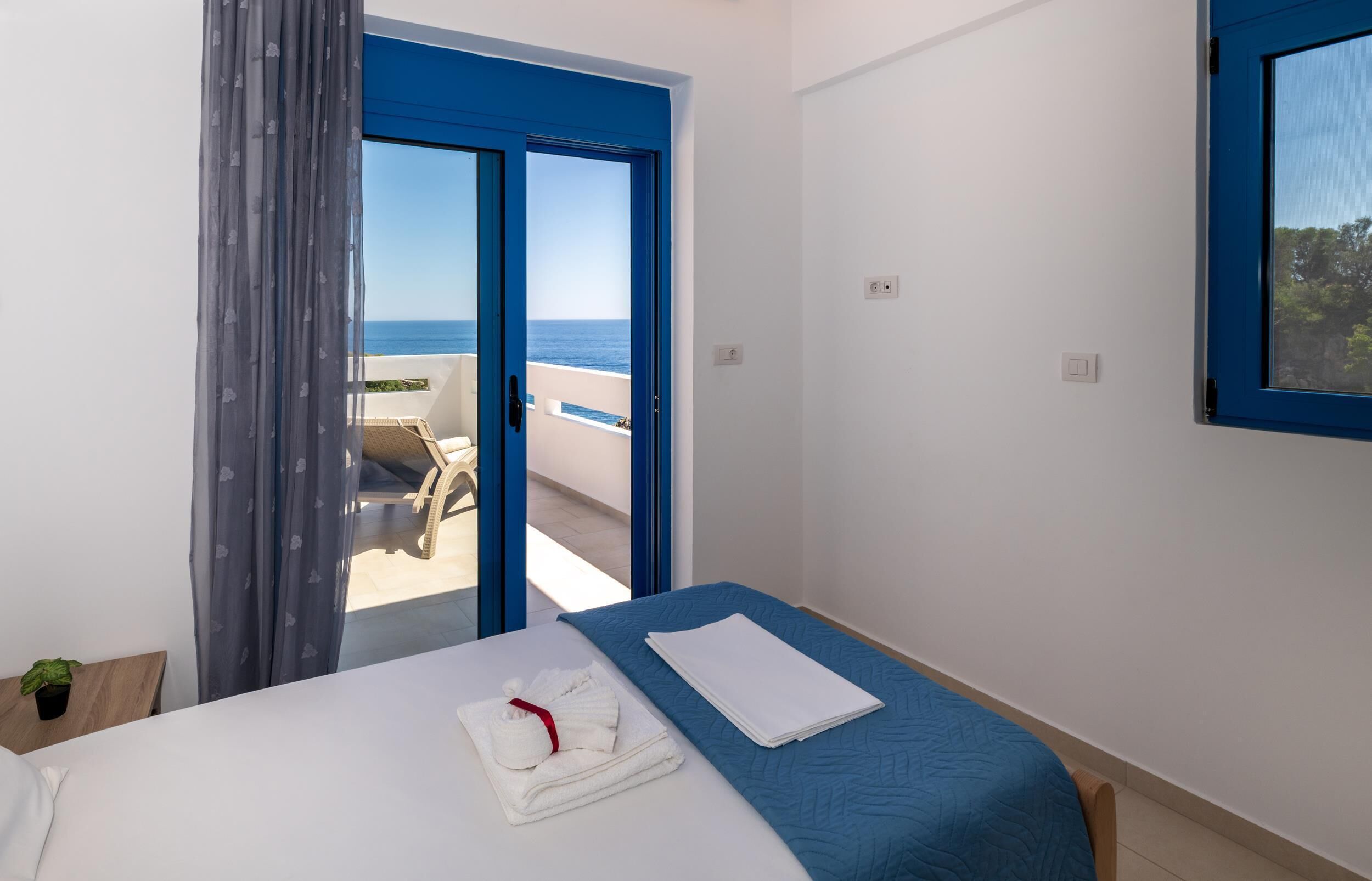 A hotel room with a bed, a blue bedspread, a white towel, and a view of the sea through a blue-framed window.