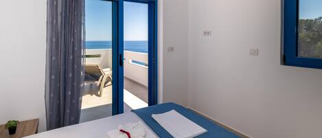 A hotel room with a bed, a blue bedspread, a white towel, and a view of the sea through a blue-framed window.