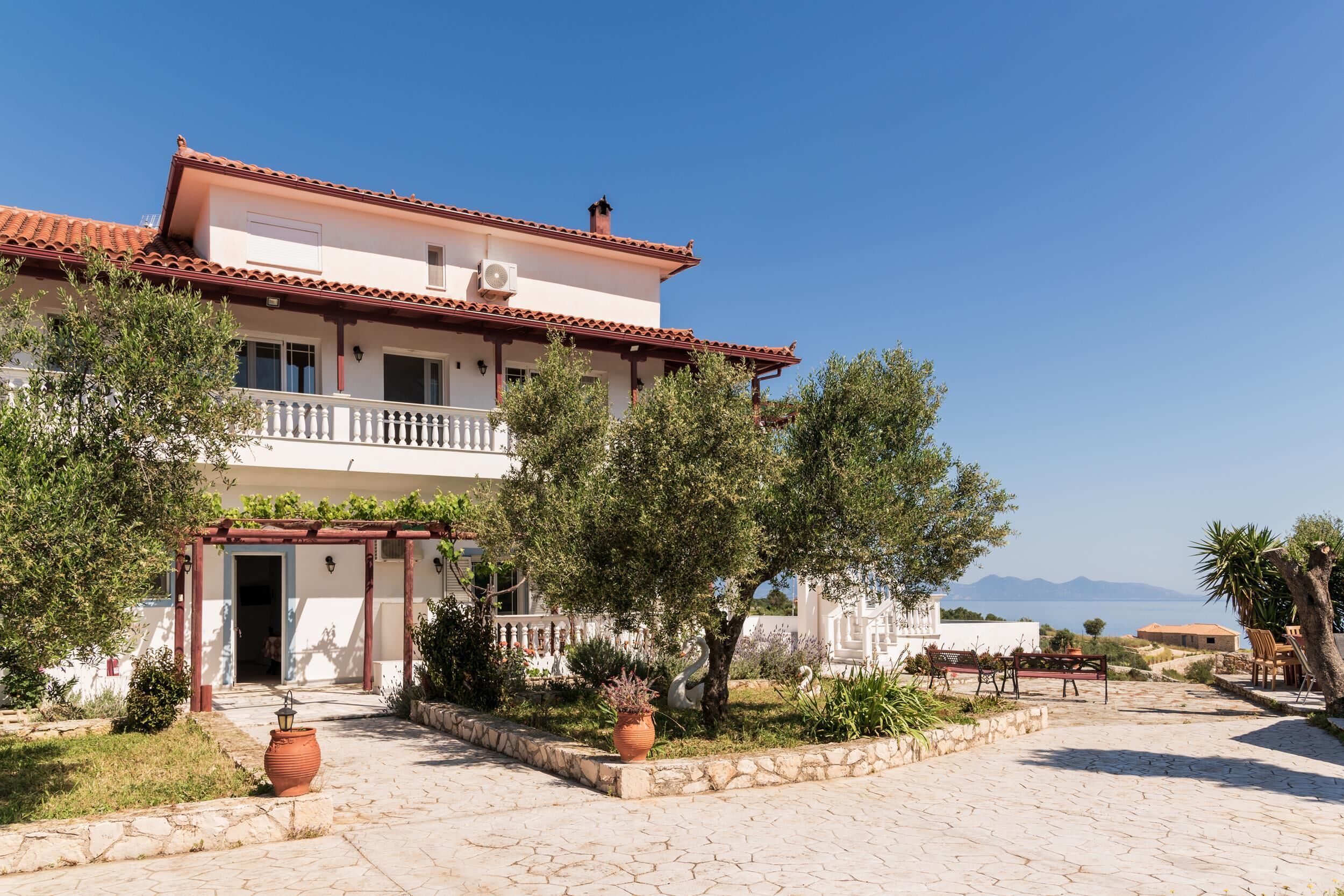 A two-story building with a balcony, a garden with potted plants, and a view of the sea.