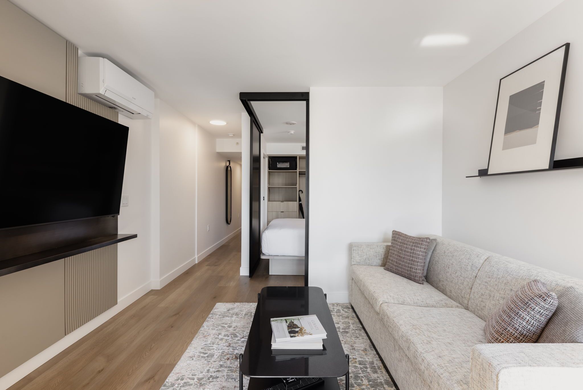A modern living room with a flat-screen TV, a sofa, a coffee table, and a hallway leading to a bedroom.