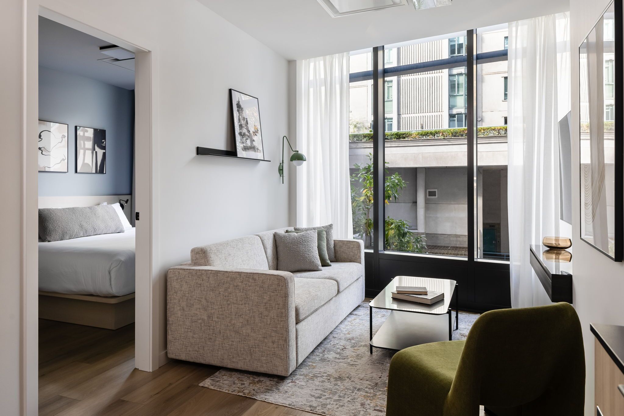 A modern living room with a sofa, a small table, and a chair. There is a bed in the adjacent room, and large windows with a view of buildings and greenery outside.