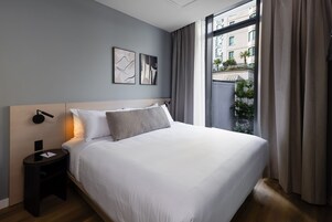 A modern hotel room with a large bed, a bedside table, and a view of the city through large windows.
