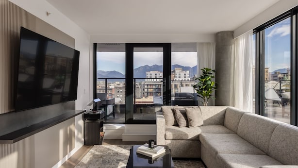 A modern living room with a large flat-screen TV, a beige sofa, a coffee table, and a view of a cityscape with mountains in the background. - Keefer House (Vancouver)