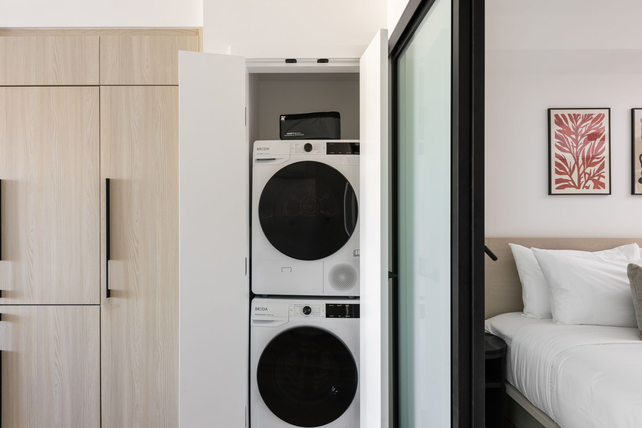A compact laundry room with two stacked washing machines, a wooden wardrobe, and a bed with white bedding.