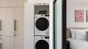A compact laundry room with two stacked washing machines, a wooden wardrobe, and a bed with white bedding.