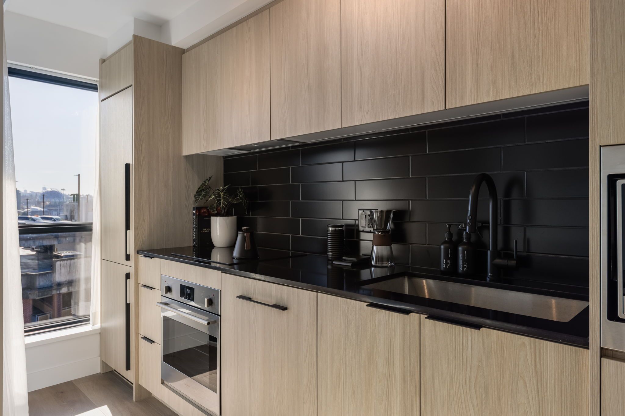 A modern kitchen with light wood cabinets, a black backsplash, stainless steel appliances, and a window overlooking a cityscape.