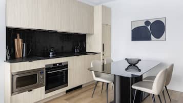 Une cuisine moderne avec des meubles en bois clair, un plan de travail noir, des appareils en acier inoxydable et une table à manger en verre, assortie de chaises blanches.