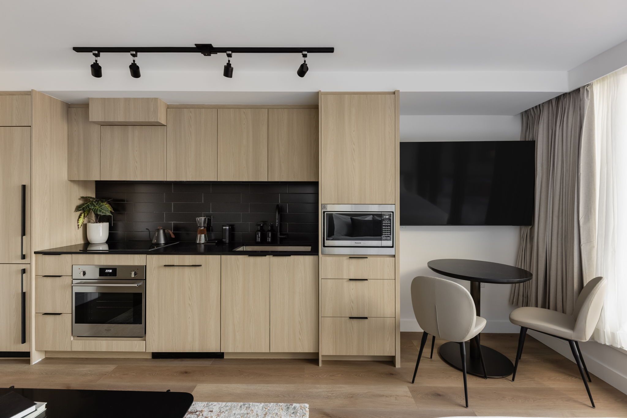 A modern kitchen with light wood cabinets, a black countertop, a microwave, and a dining area with two chairs.