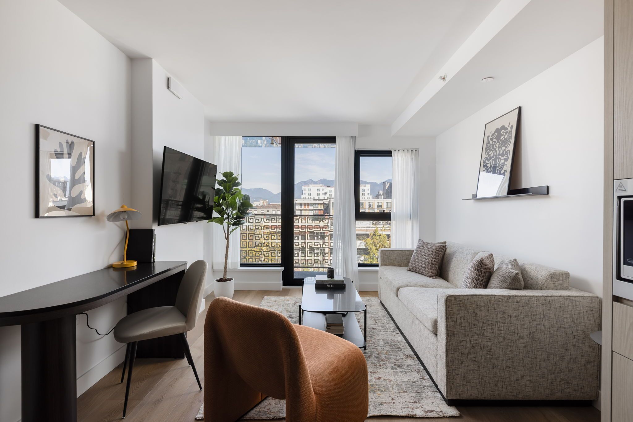 A modern living room with a sofa, a desk, and a TV. There is a large window with a view of buildings and mountains.