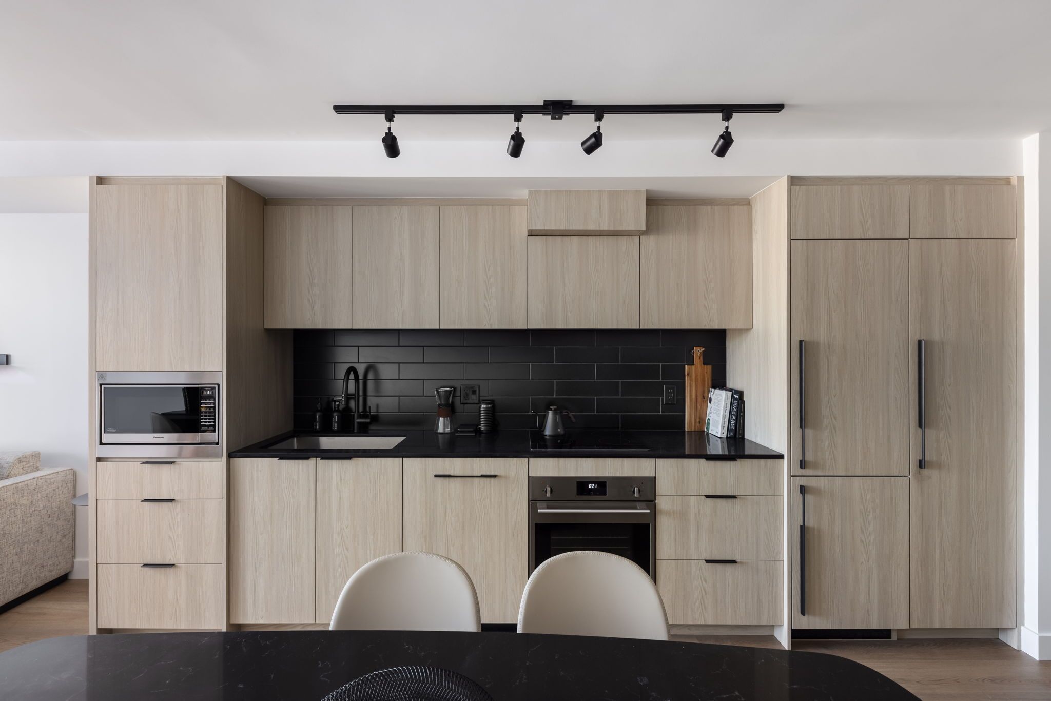 Una cocina moderna con alacenas de madera clara, encimeras negras, electrodomésticos de acero inoxidable y una mesa de comedor negra con dos sillas.