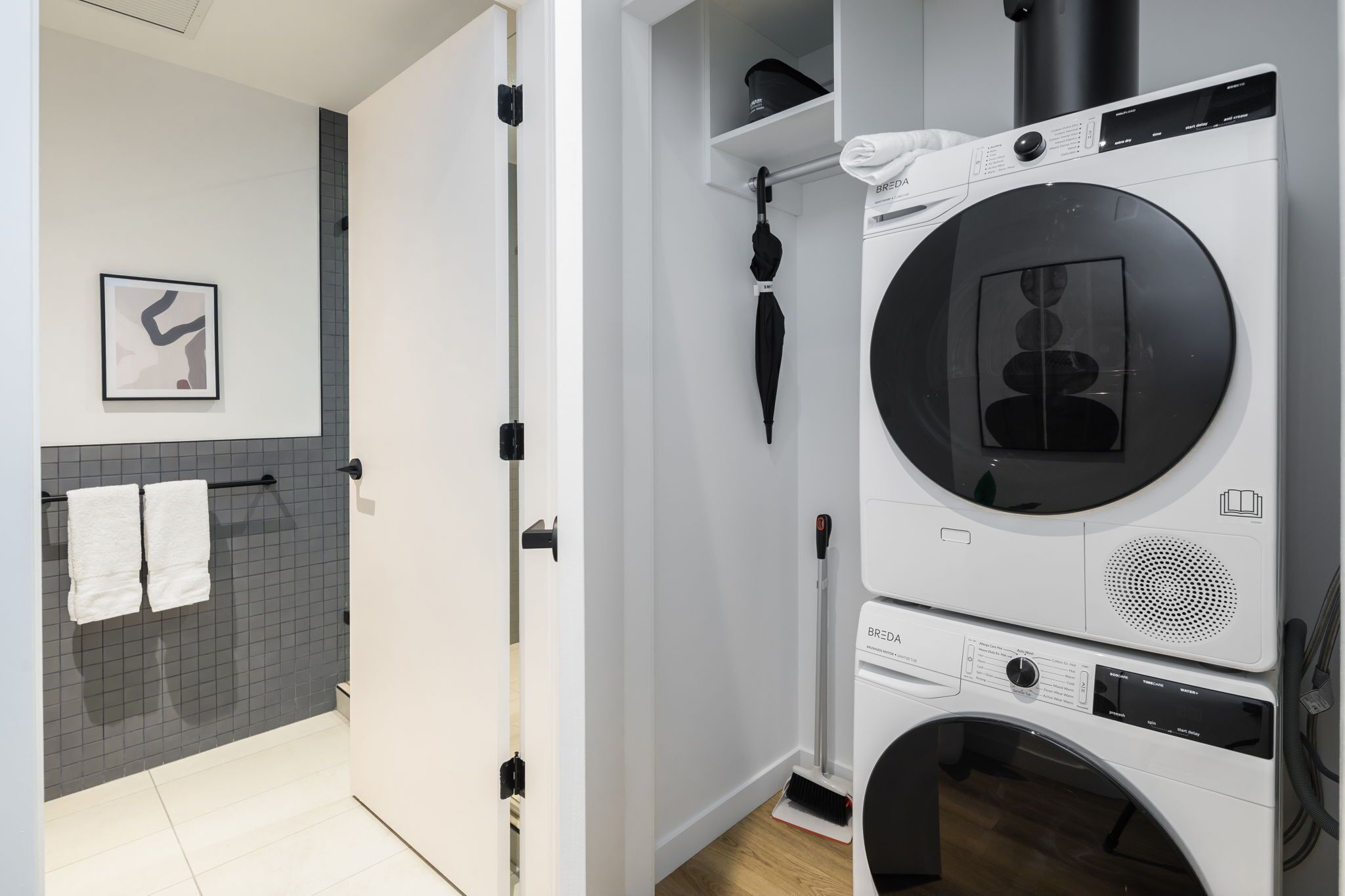 A laundry room with a front-loading washer and dryer, a towel rack with two towels, a broom, and a framed picture on the wall.