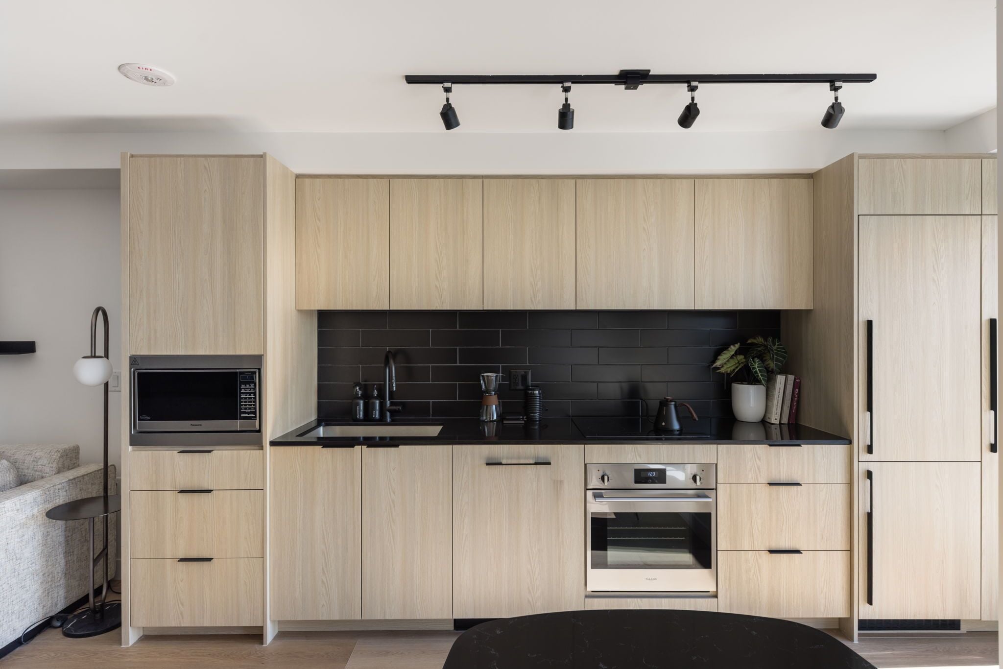 A modern kitchen with light wood cabinets, a black countertop, a microwave, an oven, and a black backsplash.
